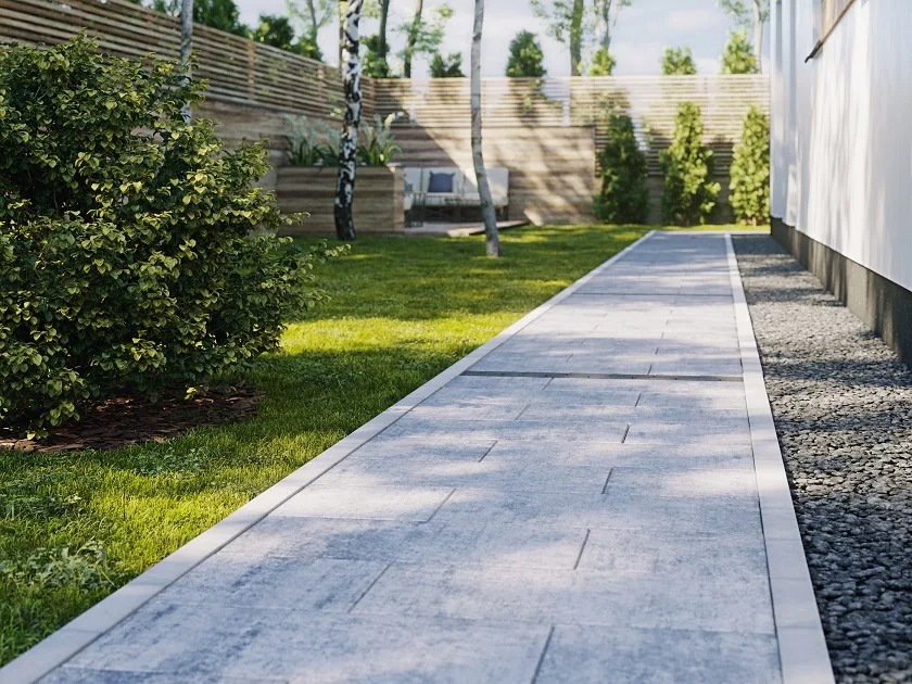 Ścieżka z jasnoszarej kostki brukowej prowadząca wzdłuż budynku, z zielonym trawnikiem i krzewami po bokach. W tle drewniany płot i drzewa.