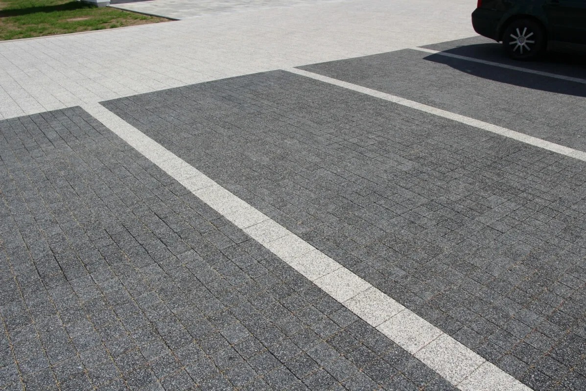 Szara kostka brukowa ułożona w pasy z białymi liniami, tworząca parking. W tle fragment samochodu i trawnik.