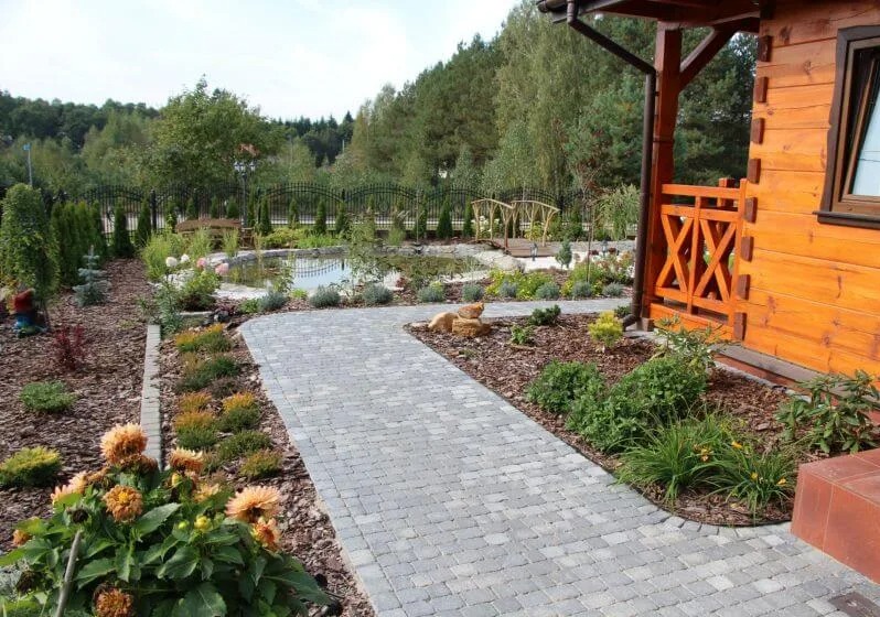 Ogród z oczkiem wodnym, ścieżką z kostki brukowej i drewnianym mostkiem. Widok z poziomu gruntu, z kwiatami na pierwszym planie i fragmentem drewnianego domu po prawej.
