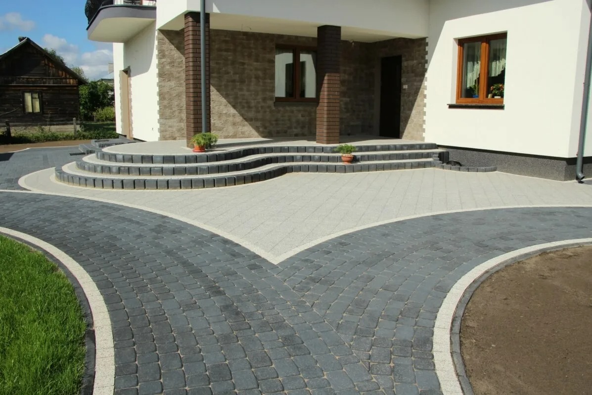 Dwukolorowa kostka brukowa tworzy rozwidlającą się ścieżkę i schody wejściowe do domu. Szara kostka kontrastuje z ciemną, a całość dopełniają rośliny w donicach.