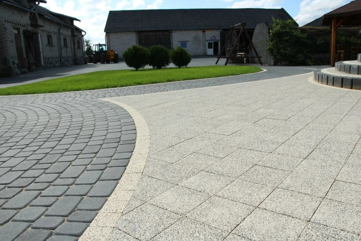 Ułożona z dwóch rodzajów kostki brukowej ścieżka na posesji. Szara kostka w łuku łączy się z jasną, kwadratową kostką. W tle budynki gospodarcze i trawnik.