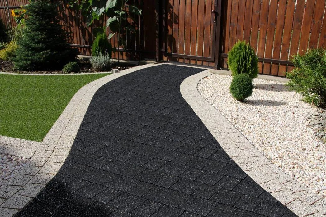 Czarny chodnik z kostki brukowej ułożony w łuk, obrzeża z jasnej kostki, obok trawnik i białe kamienie, w tle drewniany płot. Ujęcie z poziomu gruntu.