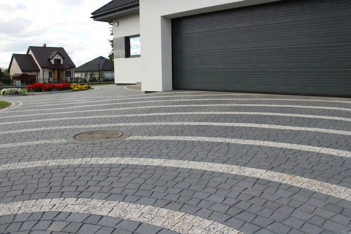 Szara kostka brukowa ułożona w łuki z białymi pasami, studzienka kanalizacyjna, brama garażowa i dom w tle. Estetyczne wykończenie podjazdu.