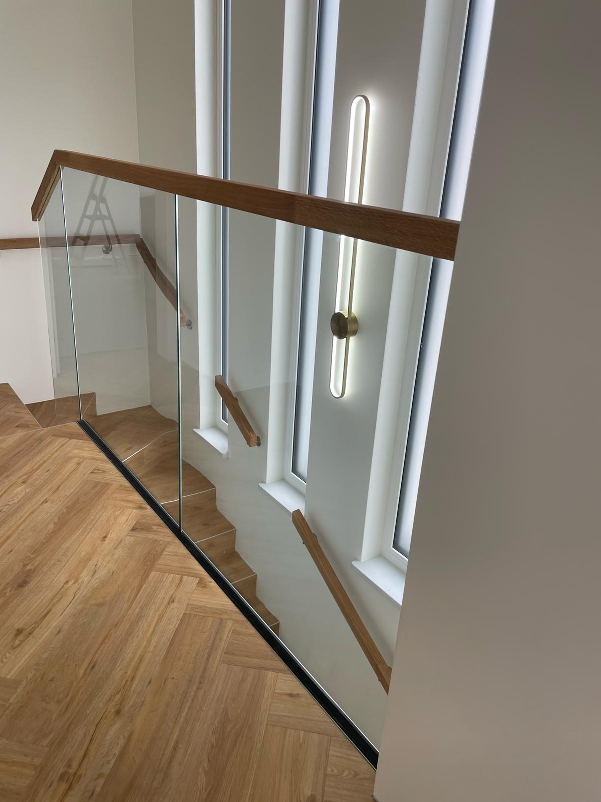 Szklana balustrada schodowa z drewnianym poręczą, widok z góry na parkiet w jodełkę i nowoczesne oświetlenie ścienne. Minimalistyczny design.