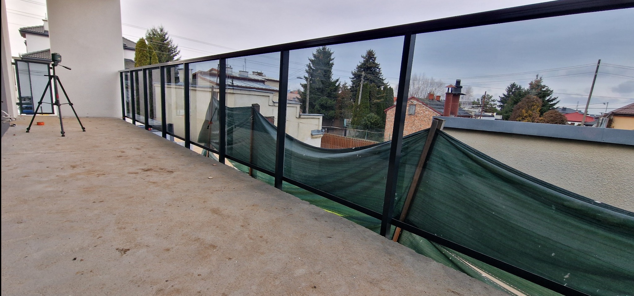 Szklana balustrada z czarnymi profilami na balkonie w trakcie montażu, widoczny niwelator laserowy na statywie i siatka zabezpieczająca. Tło stanowią budynki mieszkalne.