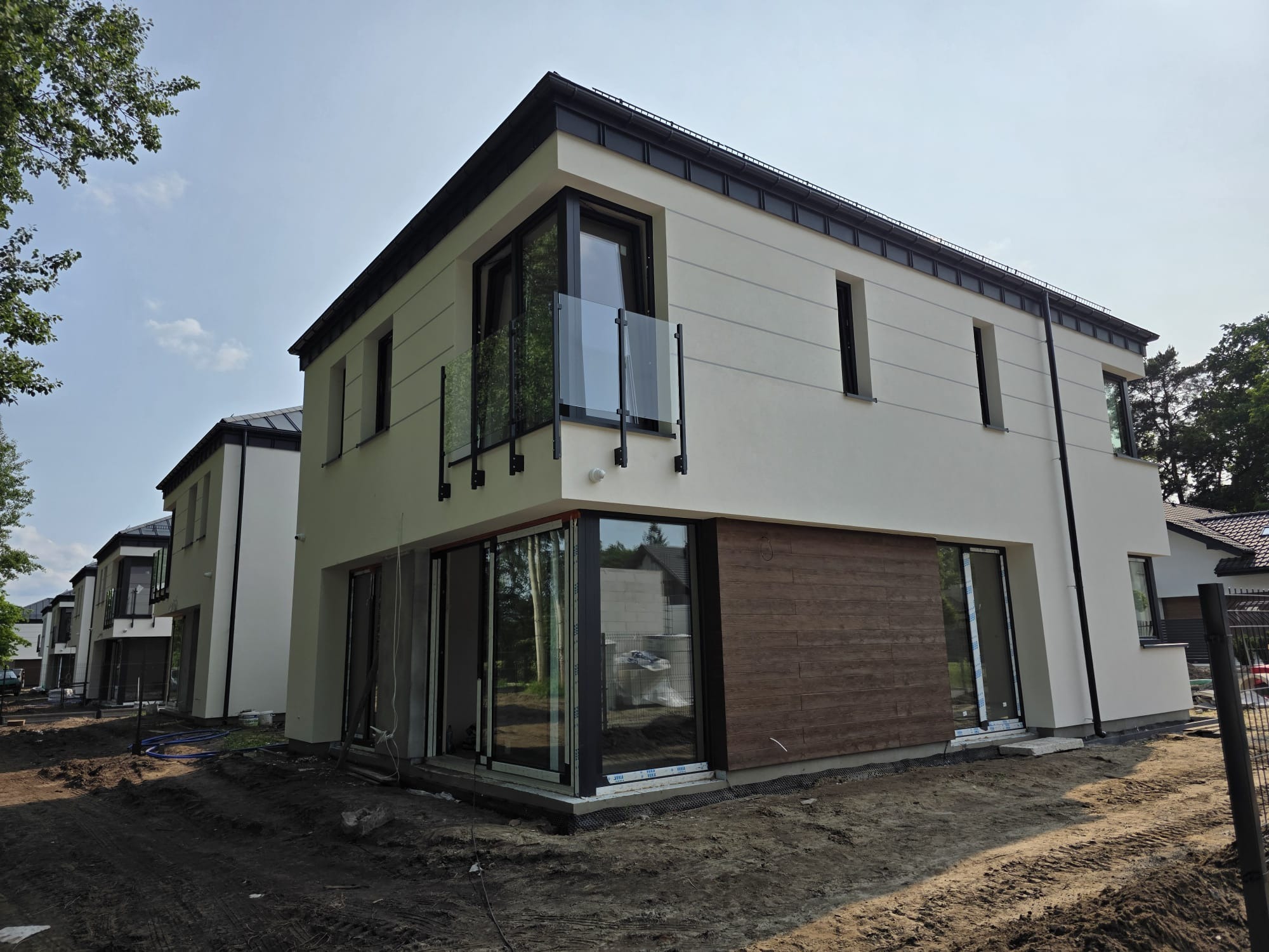 Nowoczesny dom z balkonem z balustradą szklaną i elewacją z drewnianymi panelami. Widoczne okna i drzwi balkonowe. Teren wokół domu w trakcie prac wykończeniowych.