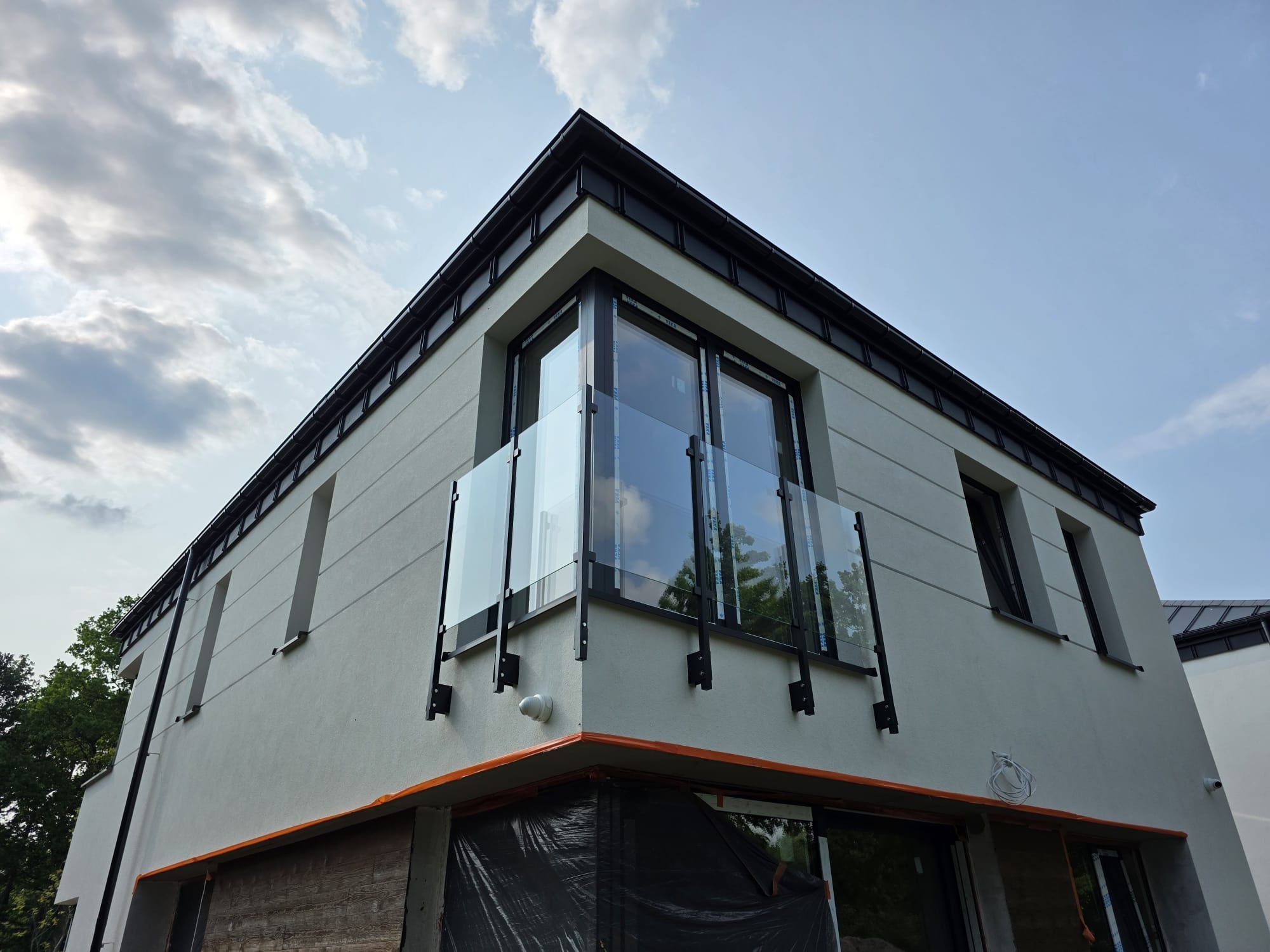 Nowoczesny budynek z balkonem zabezpieczonym szklaną balustradą z czarnymi elementami mocującymi, widziany z dołu na tle błękitnego nieba z chmurami.