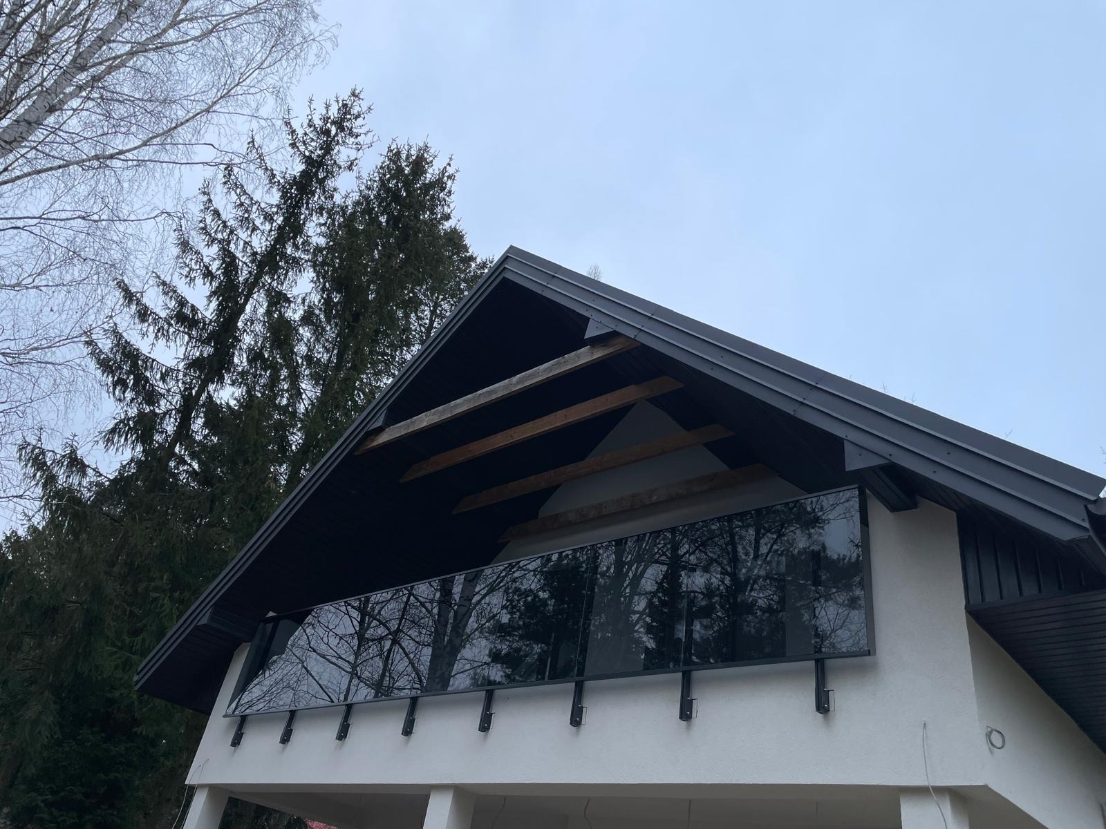 Nowoczesna balustrada szklana na balkonie domu z czarnym dachem, odbijająca drzewa. Minimalistyczny design, biała elewacja, widok z dołu.