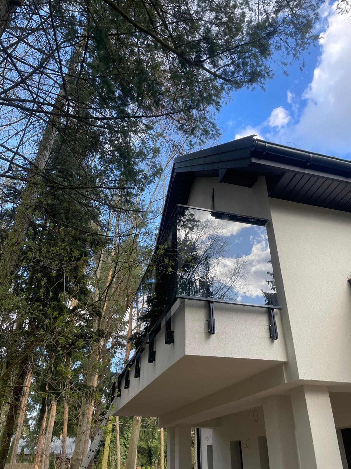 Szklana balustrada balkonowa z czarnymi elementami mocującymi, odbijająca błękitne niebo i drzewa, na tle jasnej elewacji budynku i drzew.