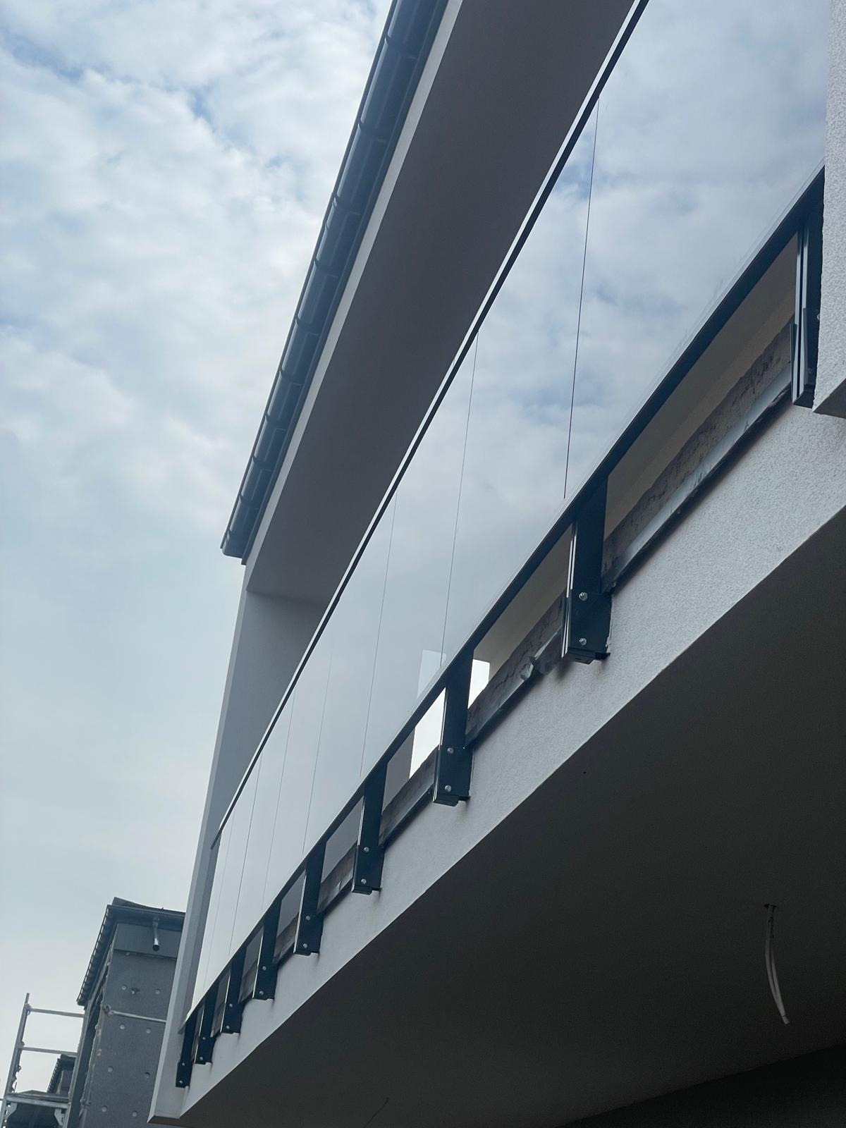 Nowoczesna balustrada szklana z czarnymi elementami mocującymi, zamontowana na balkonie budynku. Widok z dołu, pochmurne niebo w tle.