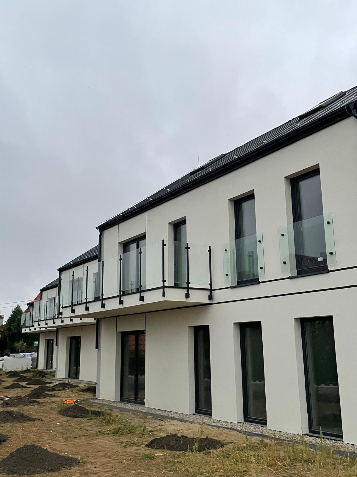 Nowoczesny budynek wielorodzinny z balkonami zabezpieczonymi szklanymi balustradami z czarnymi elementami konstrukcyjnymi. Widok na elewację i teren przed budynkiem.