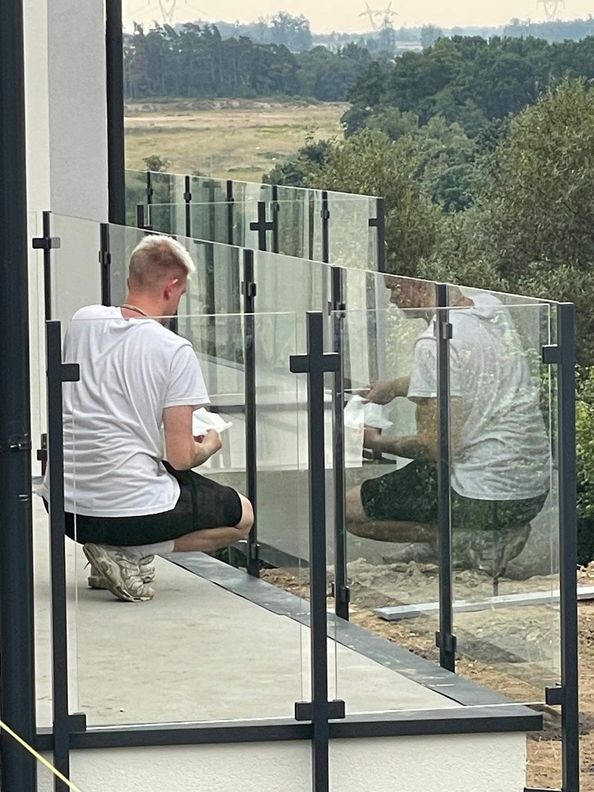 Montaż balustrady szklanej z czarnymi profilami na tarasie. Dwóch mężczyzn czyści szkło. W tle zieleń i elementy krajobrazu. Widok z zewnątrz.