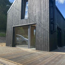 Stolarnia u Sąsiada Sławomir Dawidowicz - Czarny, nowoczesny dom z drewnianą elewacją i dużymi oknami, otoczony drewnianym tarasem. Widoczne przewody elektryczne na zewnątrz budynku.