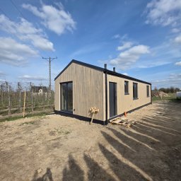 Stolarnia u Sąsiada Sławomir Dawidowicz - Gotowy, parterowy dom modułowy z jasną elewacją drewnianą i ciemnymi oknami, umieszczony na działce z widocznymi śladami budowy. Błękitne niebo z chmurami w tle.