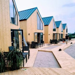 Stolarnia u Sąsiada Sławomir Dawidowicz - Rząd identycznych, drewnianych domków modułowych z niebieskimi dachami i dużymi oknami, połączonych drewnianym chodnikiem. Widoczna drabina oparta o jeden z domków.