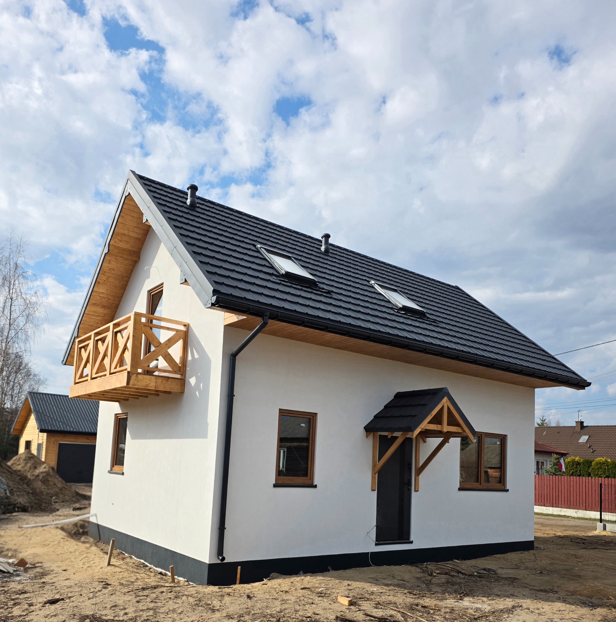 Nowoczesny dom z białymi ścianami, drewnianym balkonem i czarnym dachem. Widoczne okna dachowe i drewniane wykończenia. W tle błękitne niebo z chmurami.