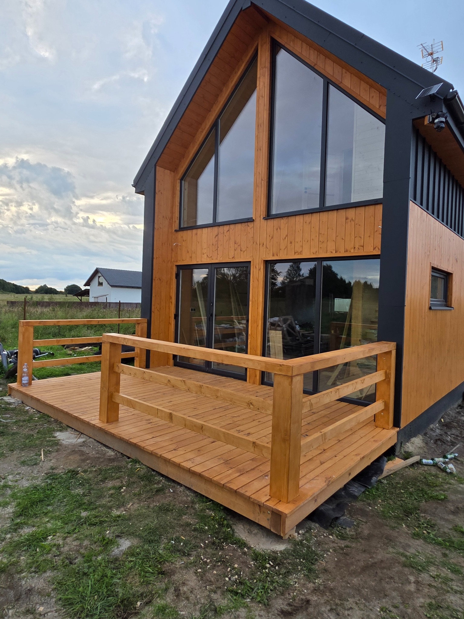 Nowoczesny dom z drewnianą elewacją i dużymi oknami, z tarasem z drewnianą balustradą. Widoczne butelki pod tarasem. W tle inny dom i pochmurne niebo.