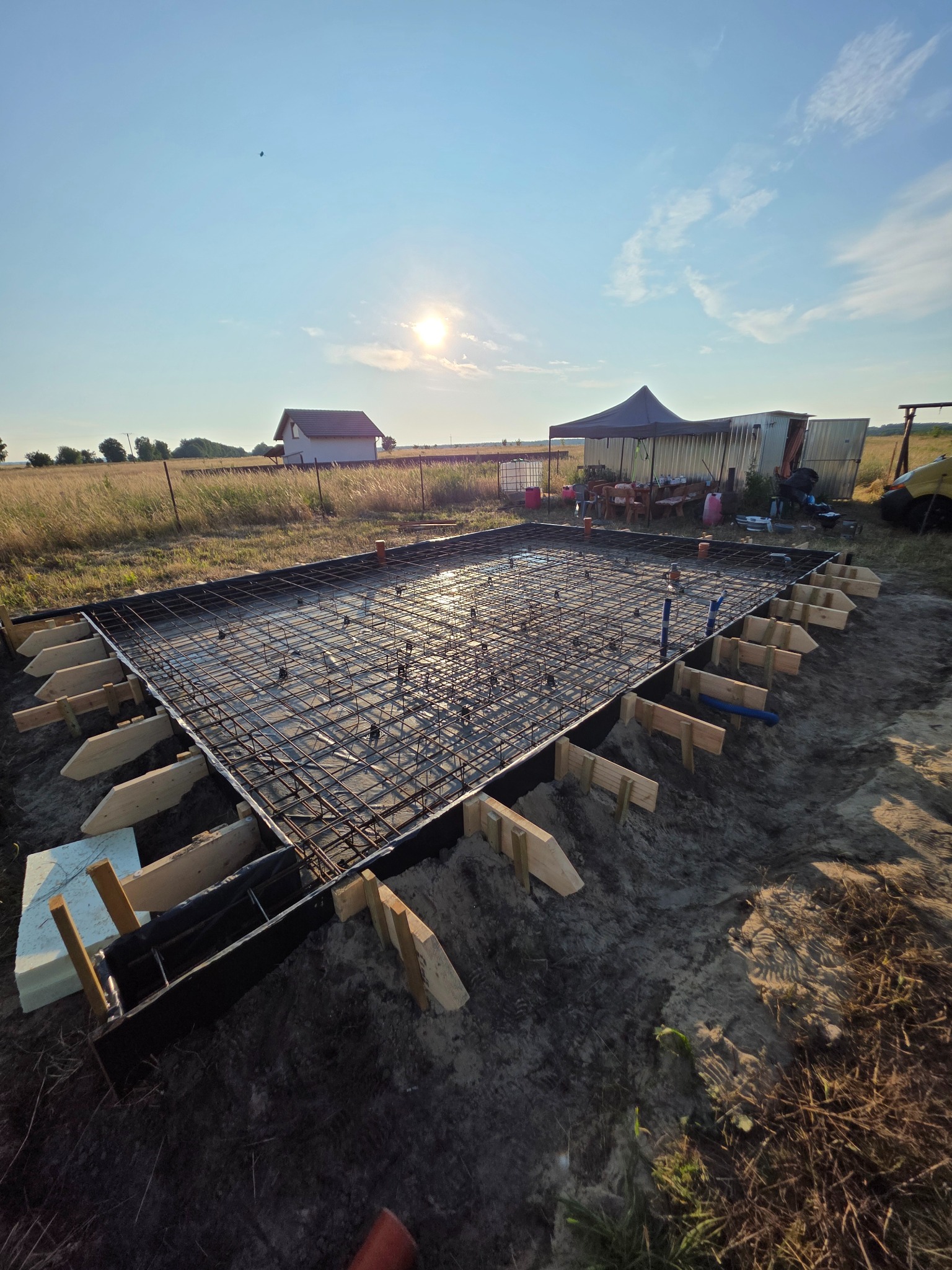 Zbrojenie fundamentu płyty, widoczne drewniane szalunki i wystające rury instalacyjne. W tle dom jednorodzinny i namiot, całość w słoneczny dzień.