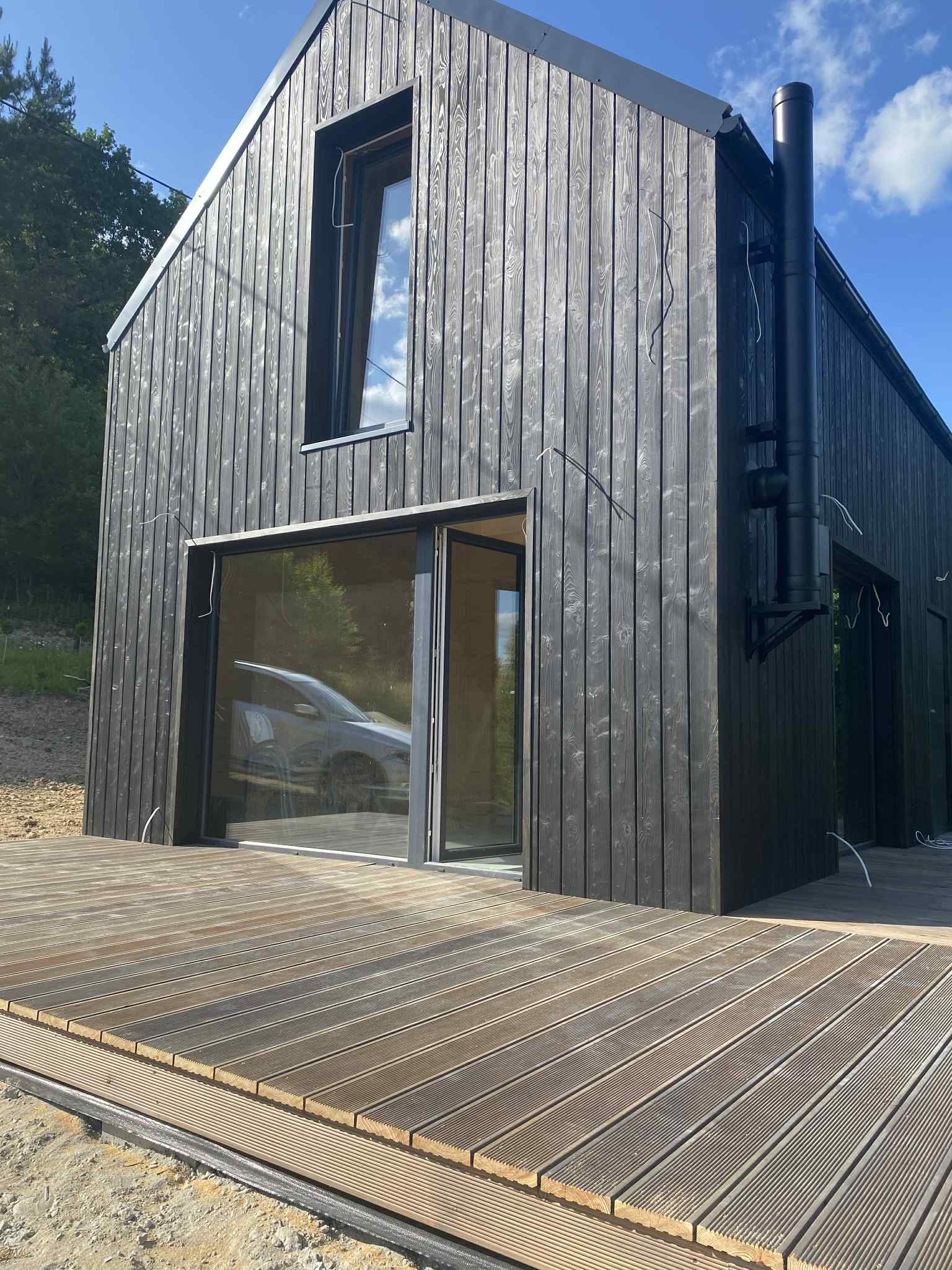 Czarny, nowoczesny dom z drewnianą elewacją i dużymi oknami, otoczony drewnianym tarasem. Widoczne przewody elektryczne na zewnątrz budynku.