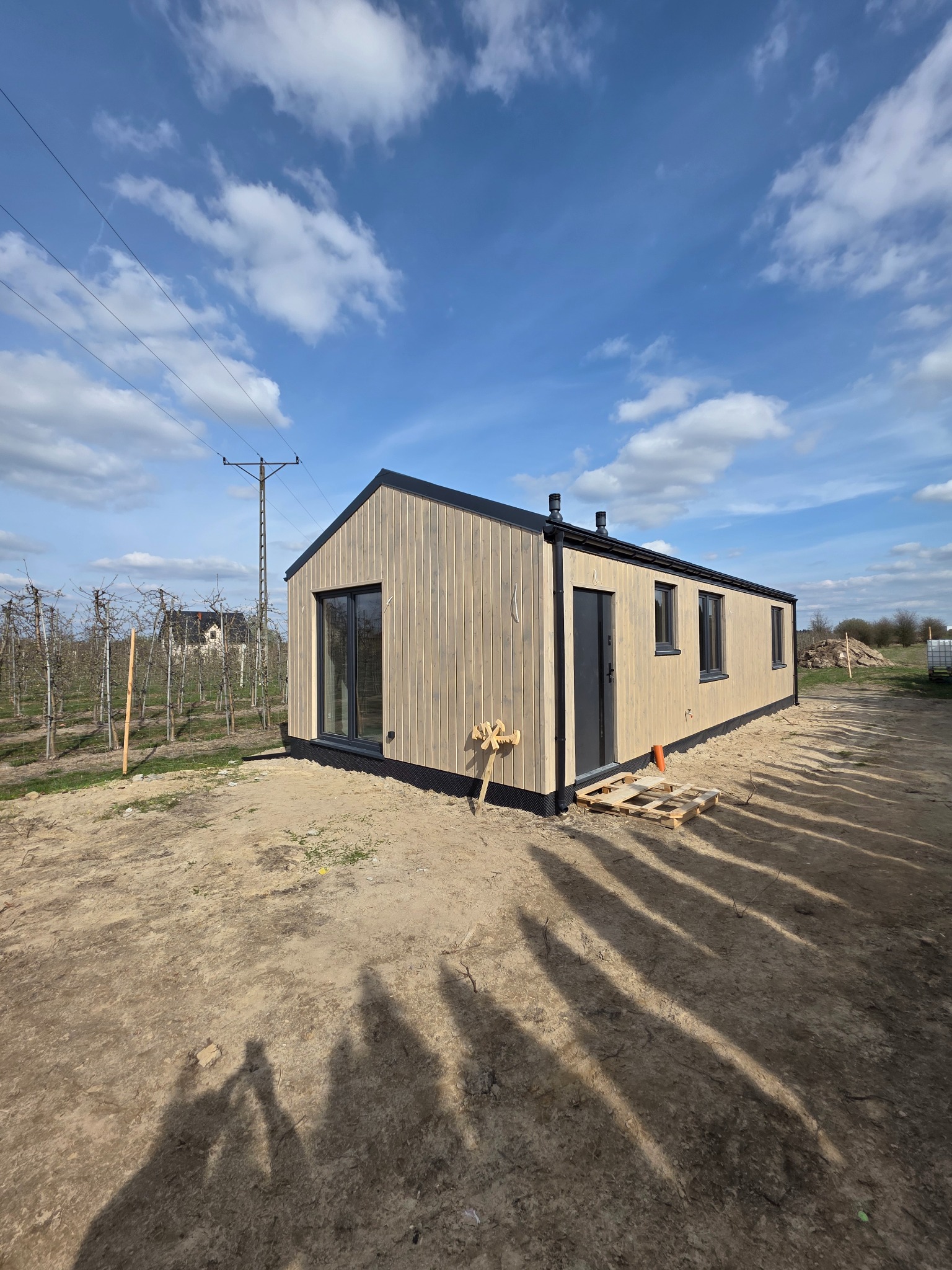 Gotowy, parterowy dom modułowy z jasną elewacją drewnianą i ciemnymi oknami, umieszczony na działce z widocznymi śladami budowy. Błękitne niebo z chmurami w tle.
