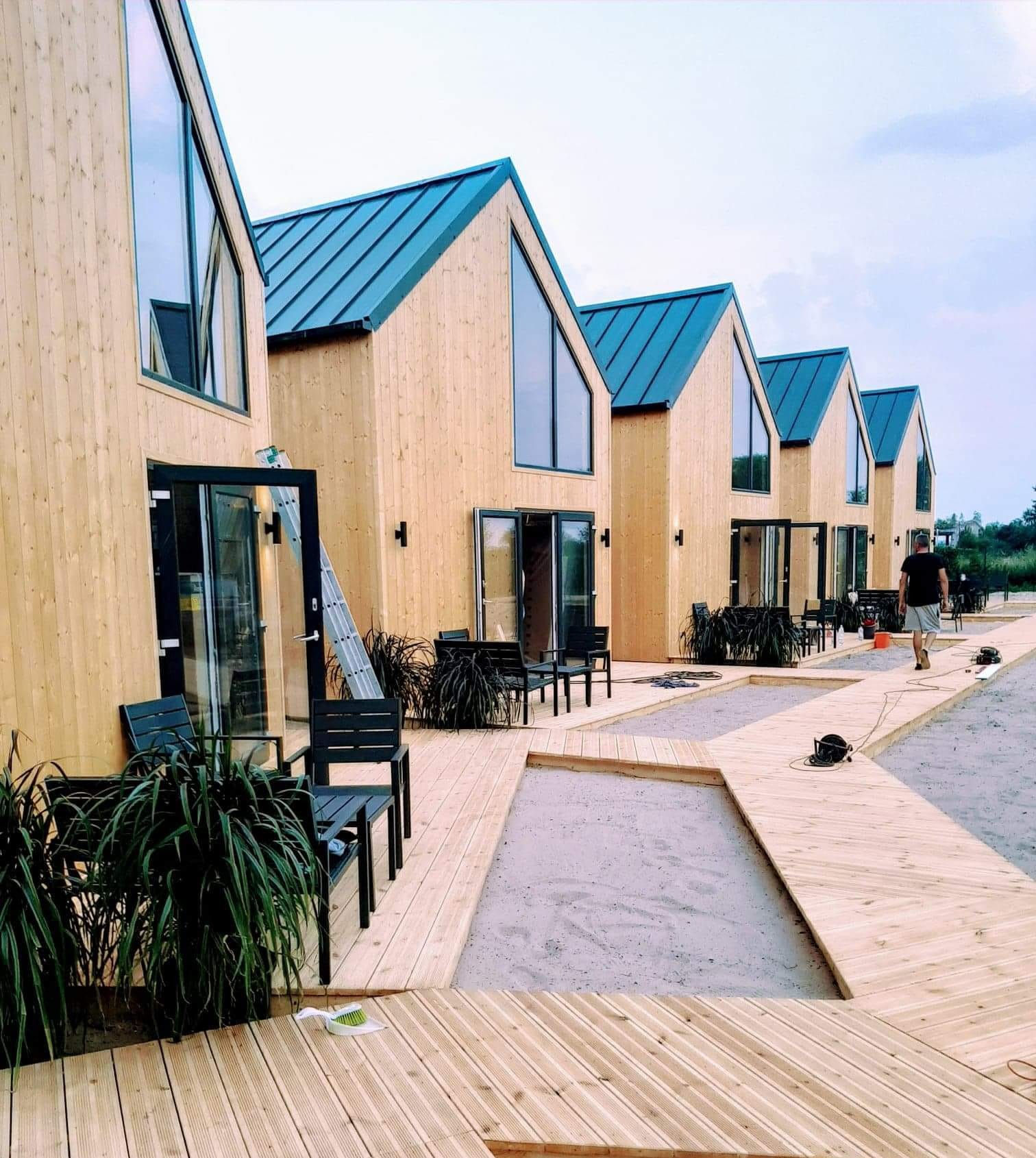 Rząd identycznych, drewnianych domków modułowych z niebieskimi dachami i dużymi oknami, połączonych drewnianym chodnikiem. Widoczna drabina oparta o jeden z domków.