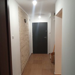 Dawid Niejodek - Korytarz po remoncie: beżowa ściana z kamienia dekoracyjnego, brązowe płytki podłogowe imitujące drewno, czarne drzwi wejściowe, białe ściany. Nowoczesne oświetlenie.
