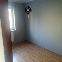 Dawid Niejodek - Pomieszczenie po malowaniu ścian na szaro, z drewnianą podłogą i nowoczesną lampą wiszącą. Widok zza otwartych drzwi, z oknem po lewej stronie.