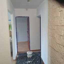 Dawid Niejodek - Białe ściany po malowaniu, zabezpieczone taśmą drzwi, wiadro farby na folii malarskiej, dekoracyjna ściana z kamienia. Widok na odnowione wnętrze mieszkania.