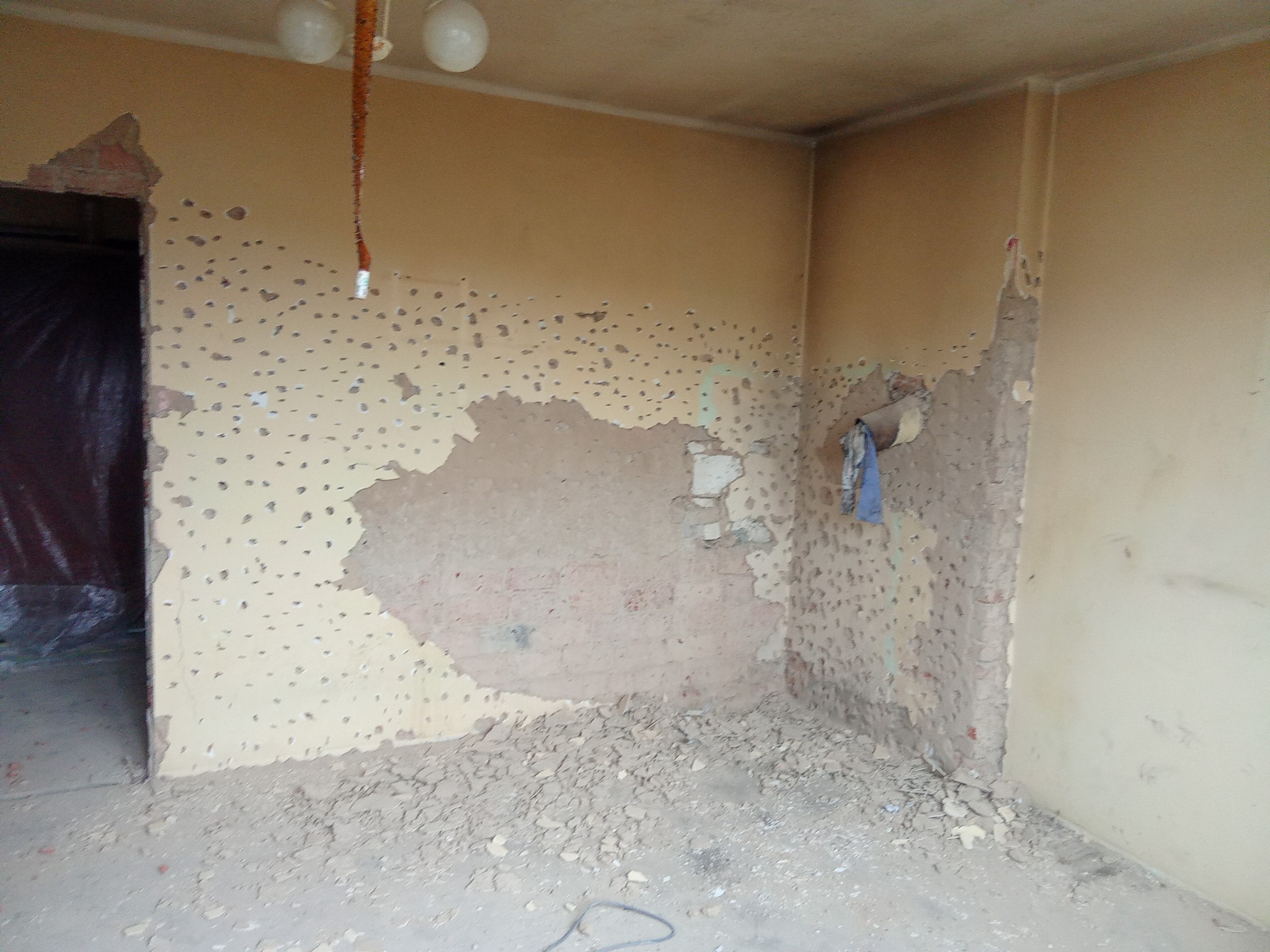 Pokój w trakcie remontu: ściany z uszkodzoną farbą, liczne dziury po kołkach, fragmenty odpadającego tynku i gruzu na podłodze. Widoczne instalacje elektryczne.