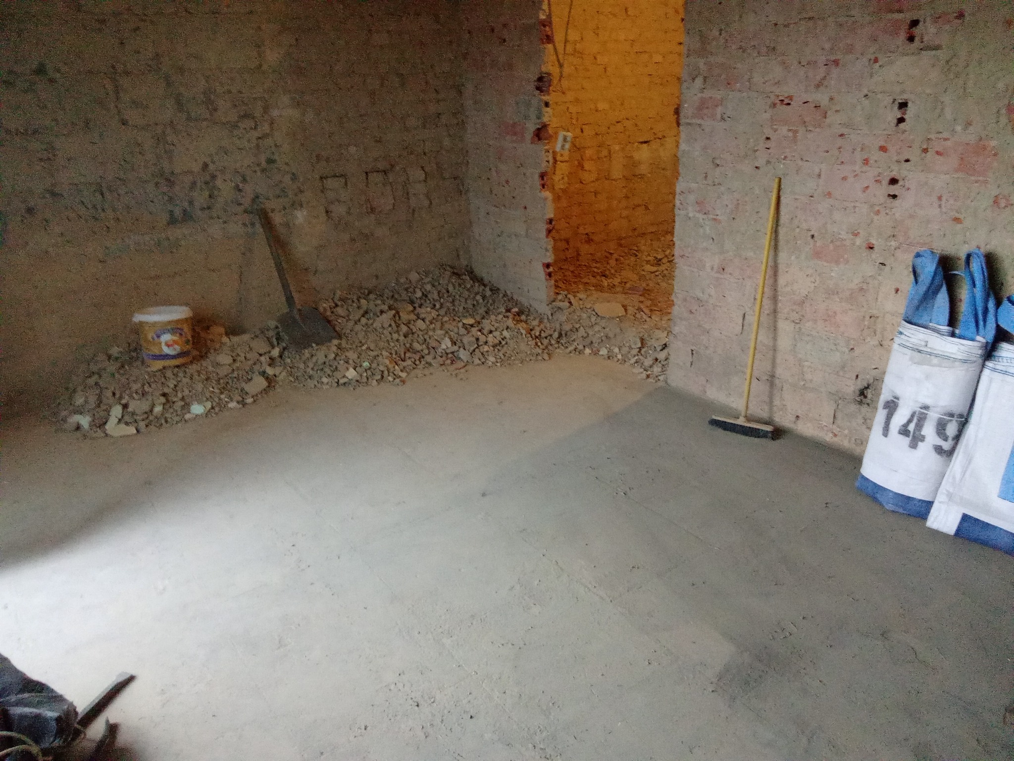 Pokój po wylewce, widoczne worki z zaprawą, resztki gruzu, łopata i miotła. Surowe ściany z cegły. Przygotowanie do dalszych prac wykończeniowych.