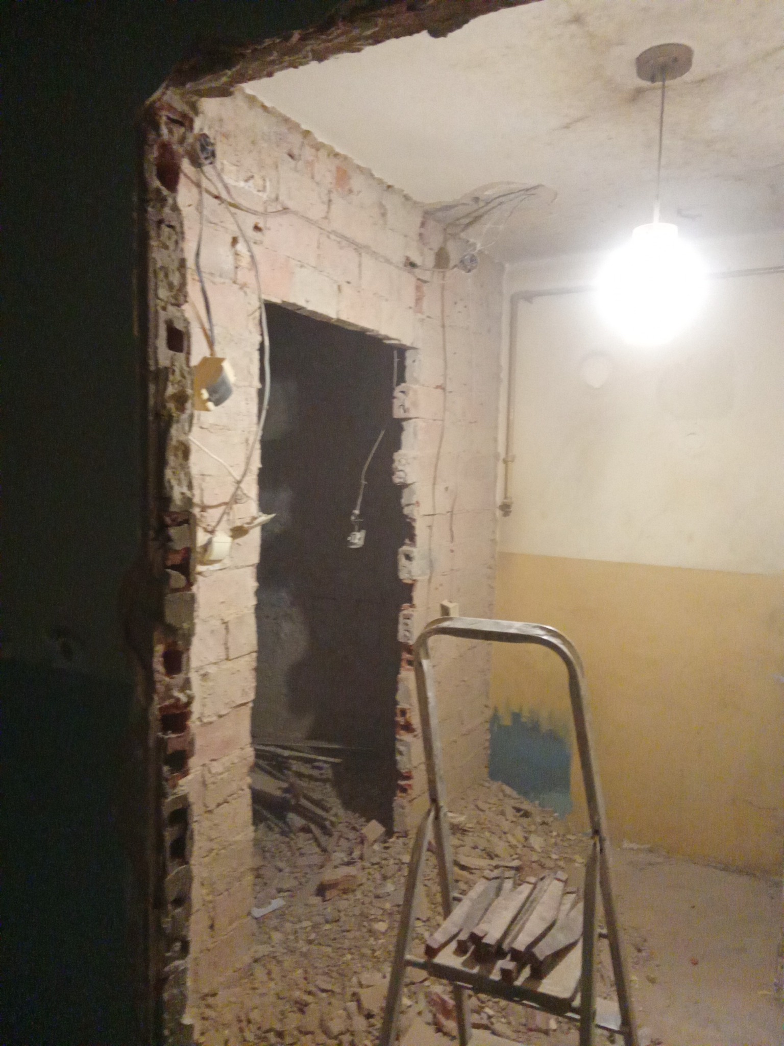 Demontaż ściany z cegły, widoczne przewody elektryczne, gruz i drabina. Etap prac remontowych w pomieszczeniu z oświetleniem sufitowym.
