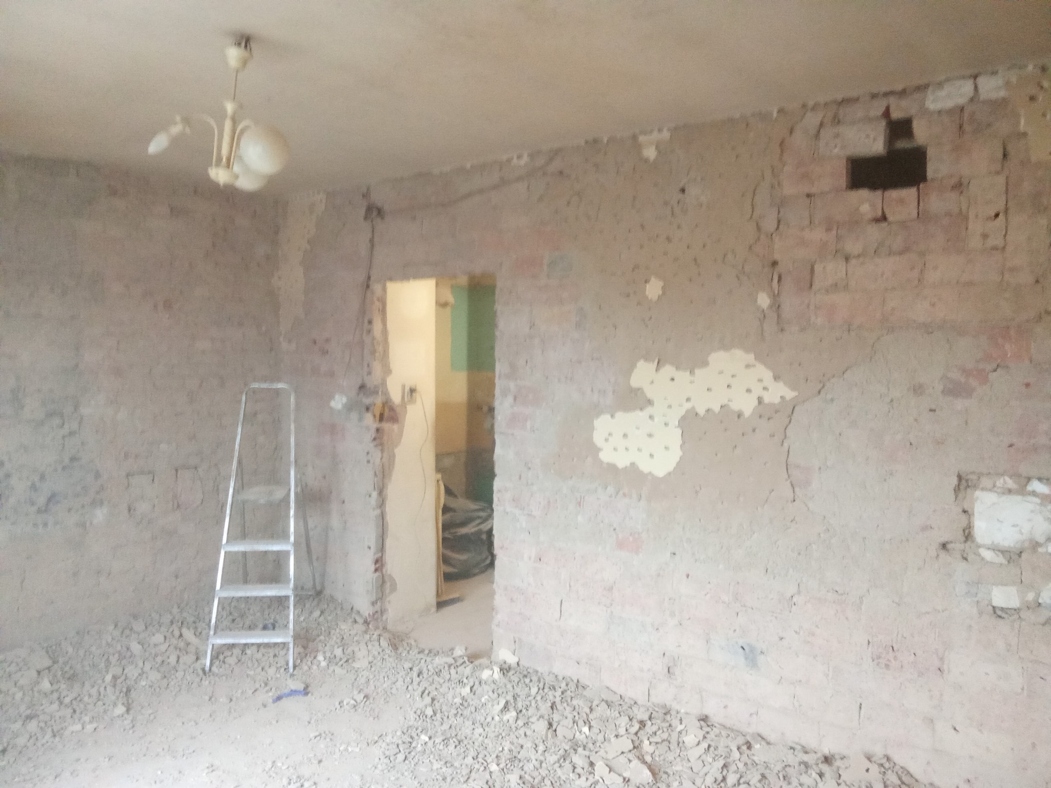 Pokój w trakcie remontu: ściany z odsłoniętą cegłą, resztki starej gładzi, drabina aluminiowa, gruz na podłodze. Widoczne ślady po instalacjach i otwór w ścianie.