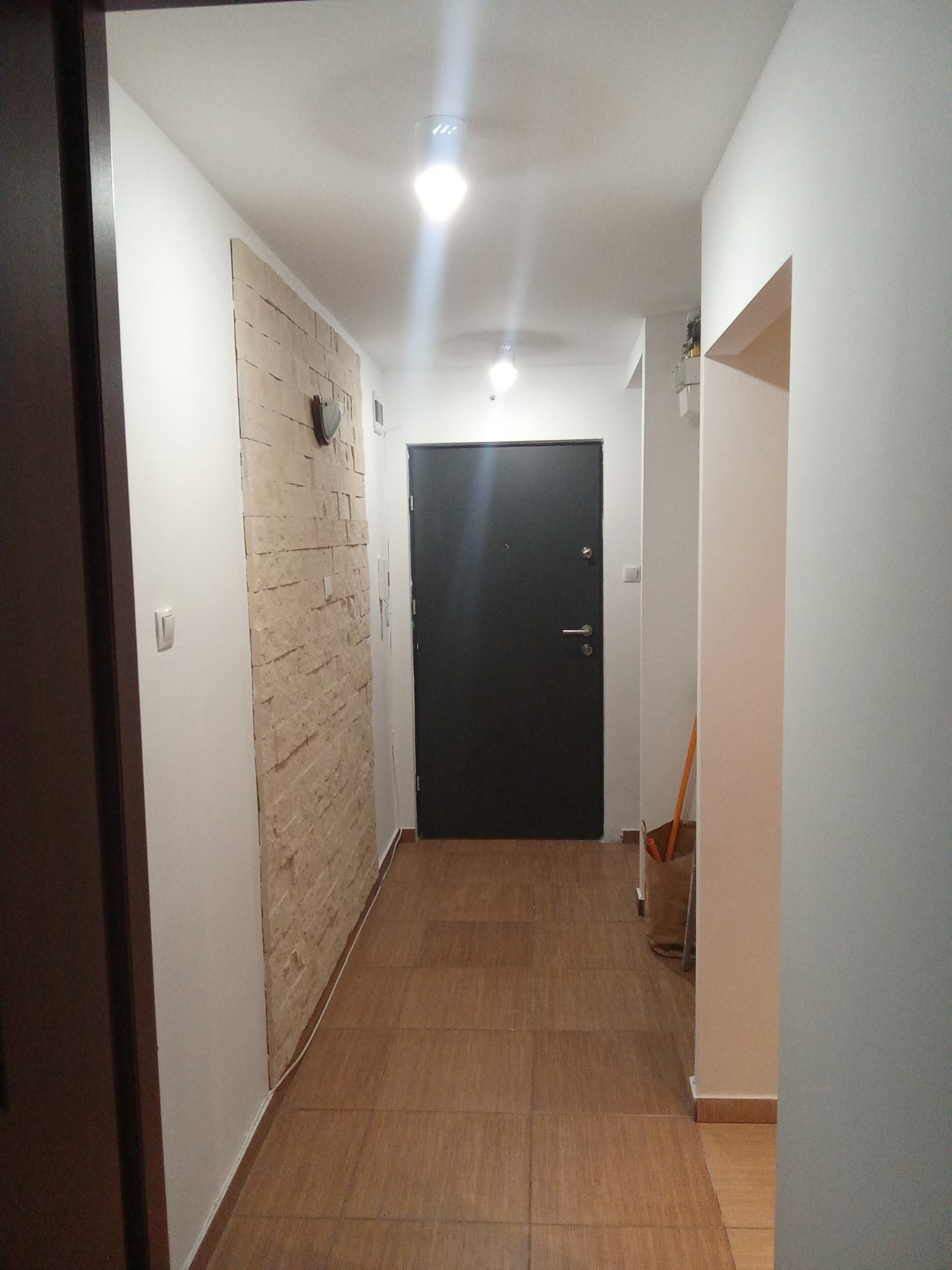 Korytarz po remoncie: beżowa ściana z kamienia dekoracyjnego, brązowe płytki podłogowe imitujące drewno, czarne drzwi wejściowe, białe ściany. Nowoczesne oświetlenie.