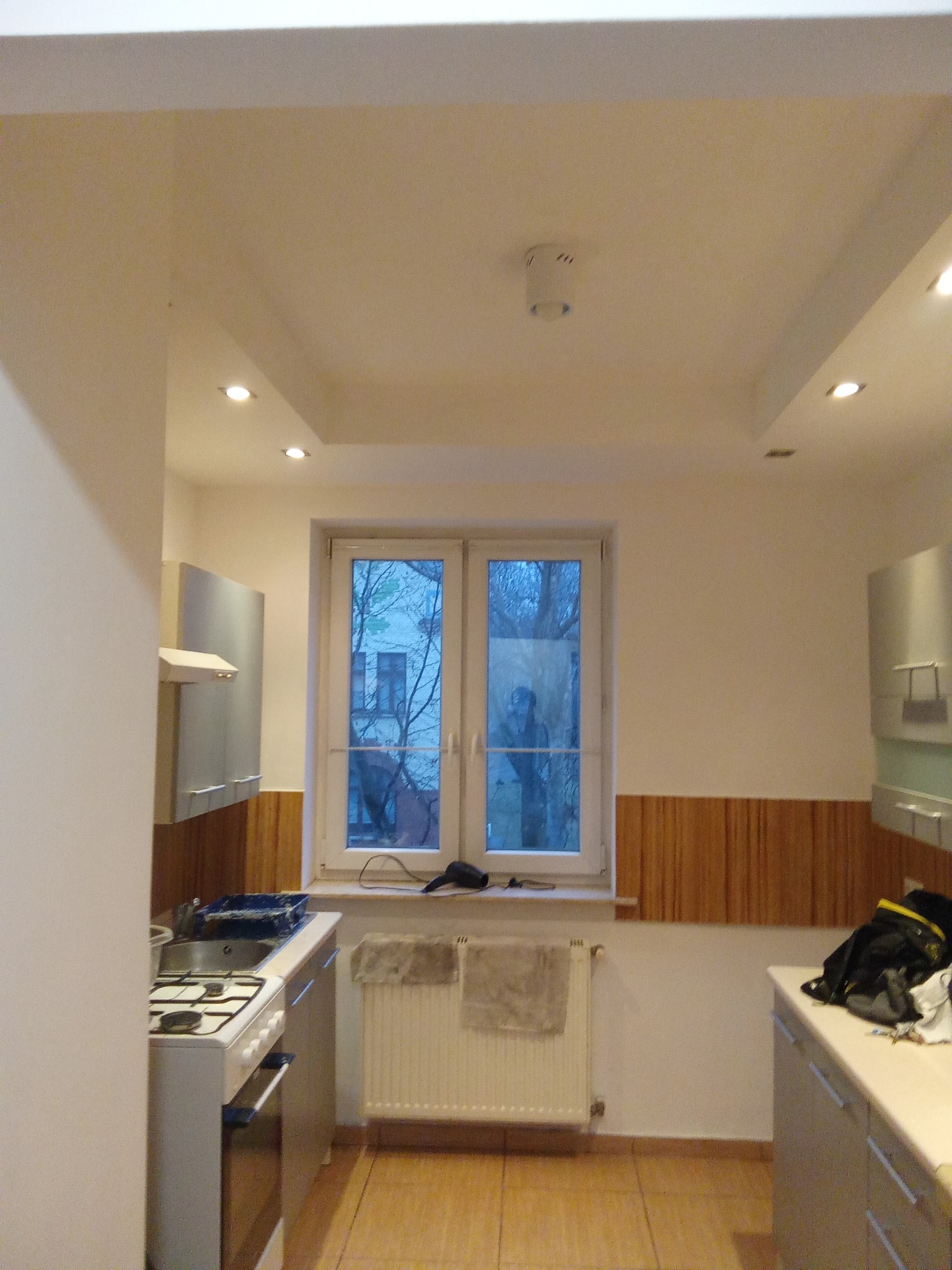 Jasna kuchnia po remoncie z białymi ścianami, drewnianą boazerią i nowoczesnymi szafkami. Widoczne okno z widokiem na drzewa i budynek. Oświetlenie sufitowe.