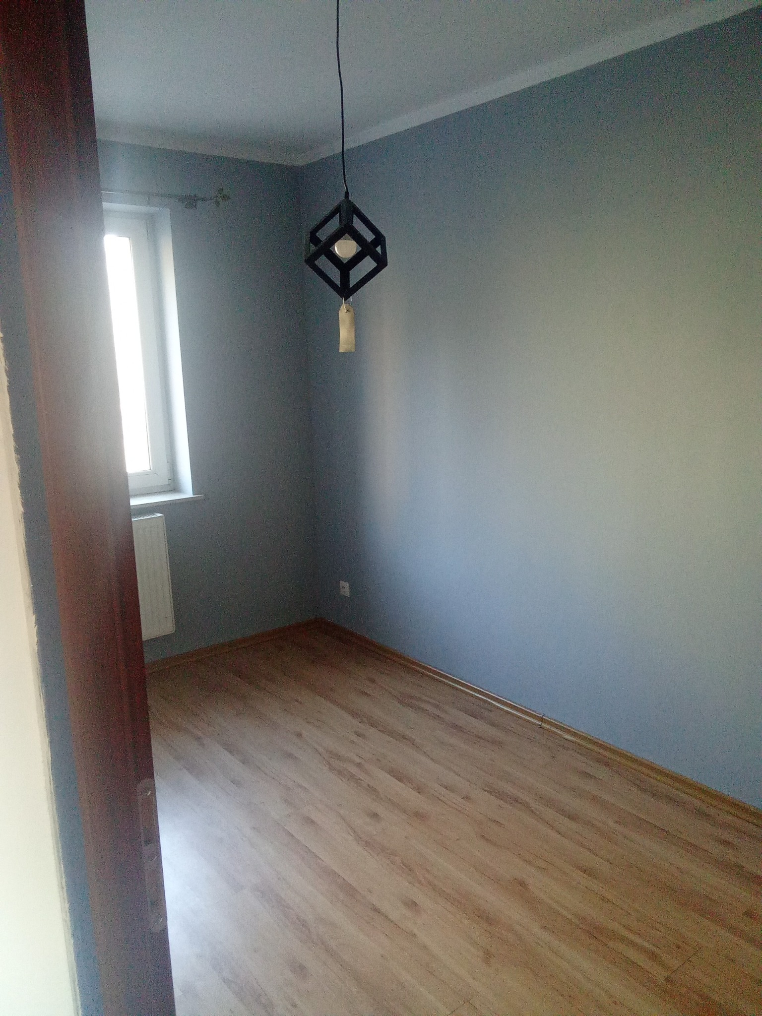 Pomieszczenie po malowaniu ścian na szaro, z drewnianą podłogą i nowoczesną lampą wiszącą. Widok zza otwartych drzwi, z oknem po lewej stronie.