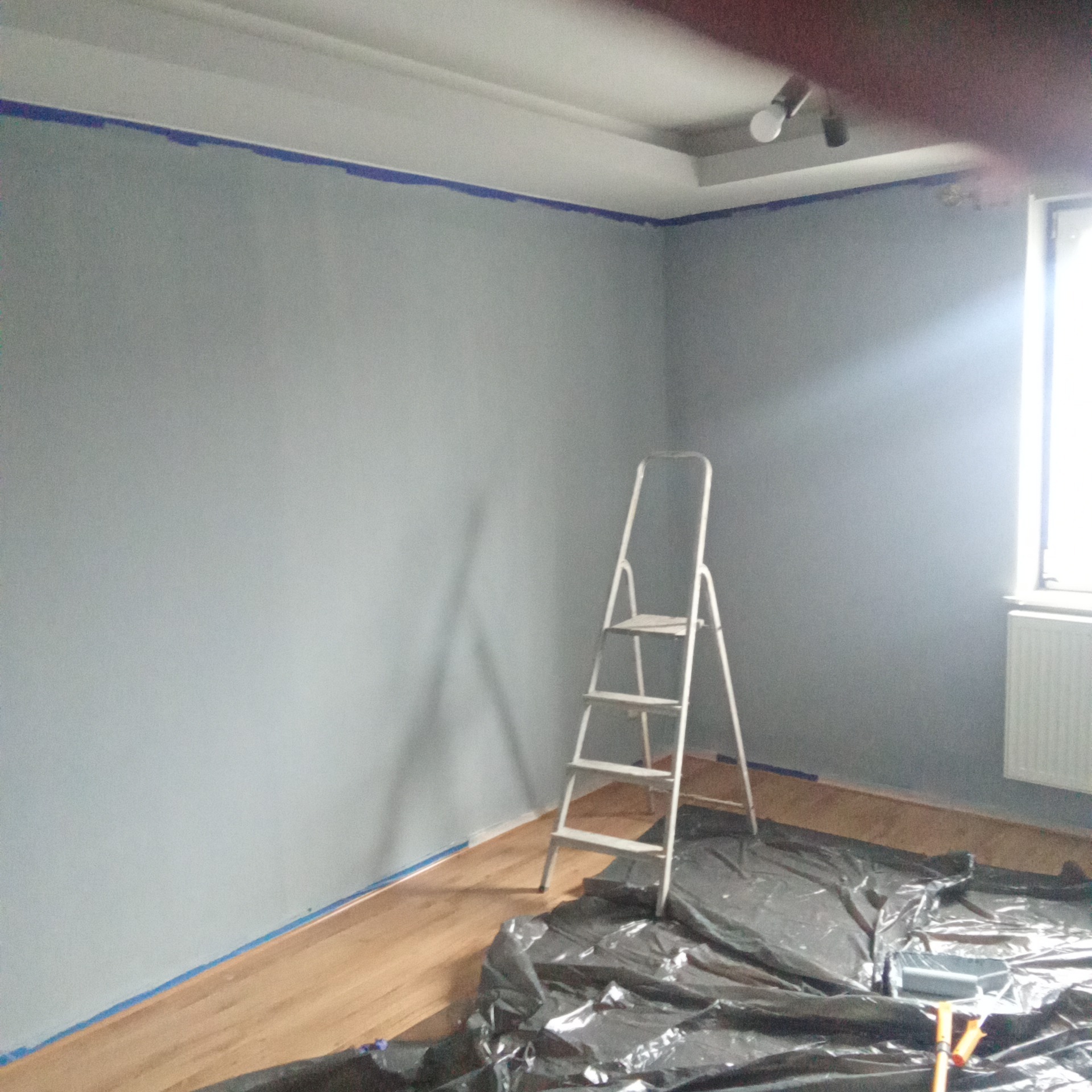 Pokój w trakcie malowania na szaro, zabezpieczony folią i taśmą malarską. Widoczna drabina malarska i akcesoria malarskie. Świeżo pomalowane ściany.