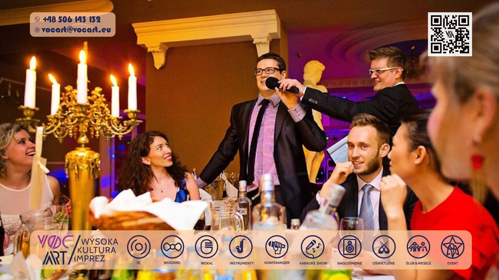 Wokalista z mikrofonem w dłoni, w otoczeniu gości przy stole, podczas występu na imprezie. W tle świecznik i dekoracje. Ujęcie z perspektywy widza.