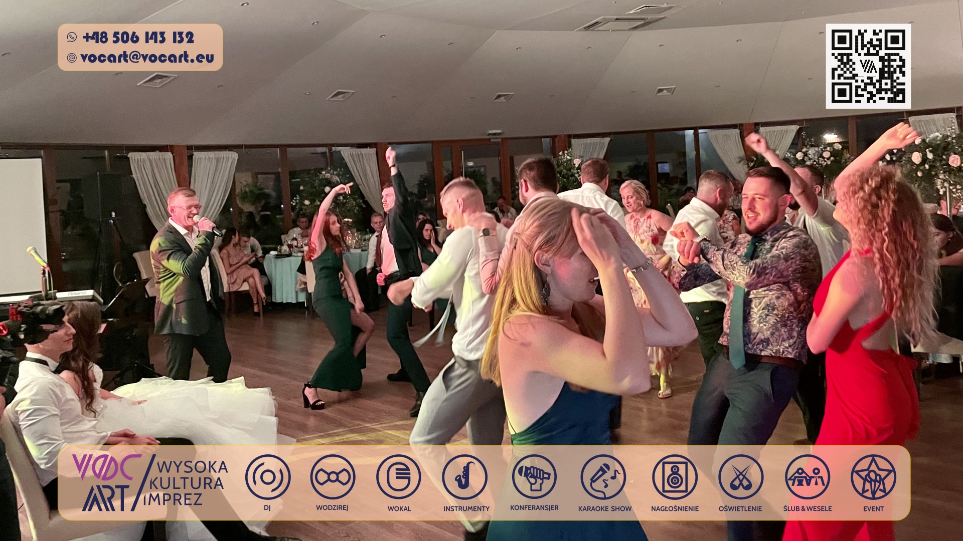 Roztańczony tłum gości weselnych, wokalista z mikrofonem na pierwszym planie, parkiet taneczny, dekoracje kwiatowe, dynamiczne ujęcie z imprezy.