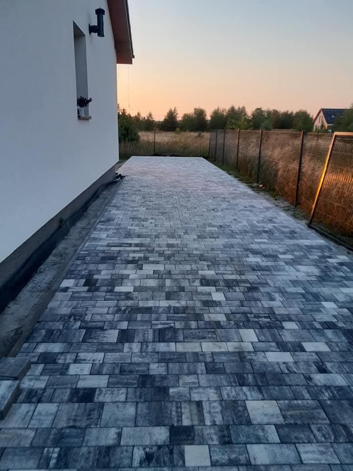 Ścieżka z szarej kostki przy domu, widok z góry, w tle zachodzące słońce. Część kostki jeszcze nieułożona, widoczny piasek. Estetyczne wykończenie.