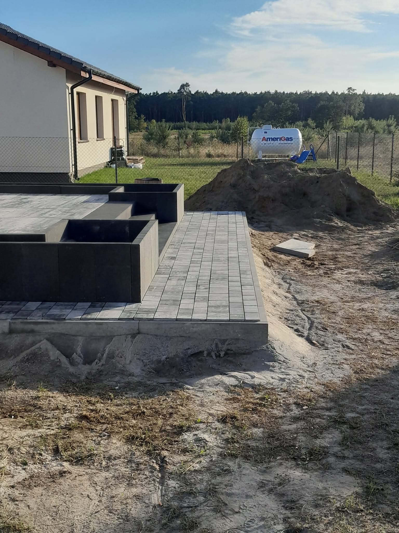 Ułożona szara kostka na podwyższeniu z donicami, w tle dom i las. Widoczny zbiornik na gaz. Prace brukarskie w toku, teren częściowo przygotowany.