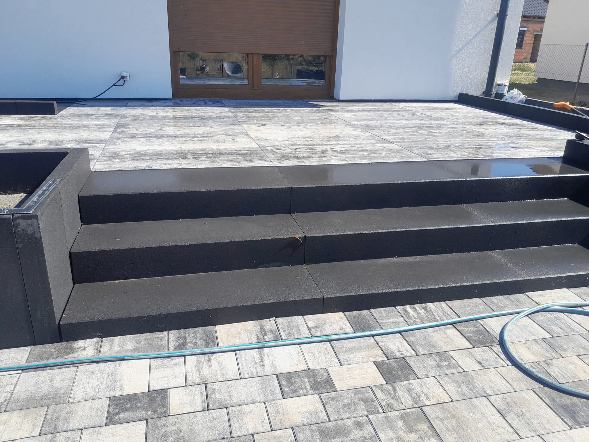 Nowoczesne schody z ciemnej kostki prowadzące na taras z jasnoszarej kostki. Widoczny fragment węża ogrodowego i elewacja budynku w tle.