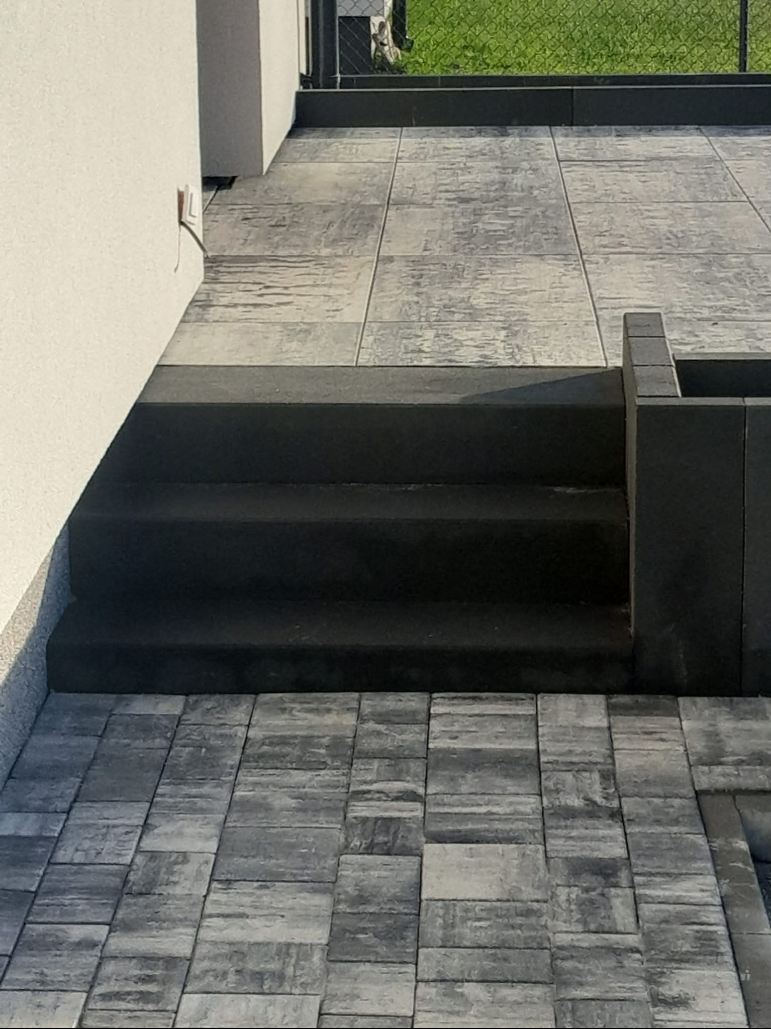 Schody i taras wyłożone szarą kostką o nieregularnej fakturze, z widocznym fragmentem ściany budynku i zieleni w tle. Nowoczesne wykończenie.