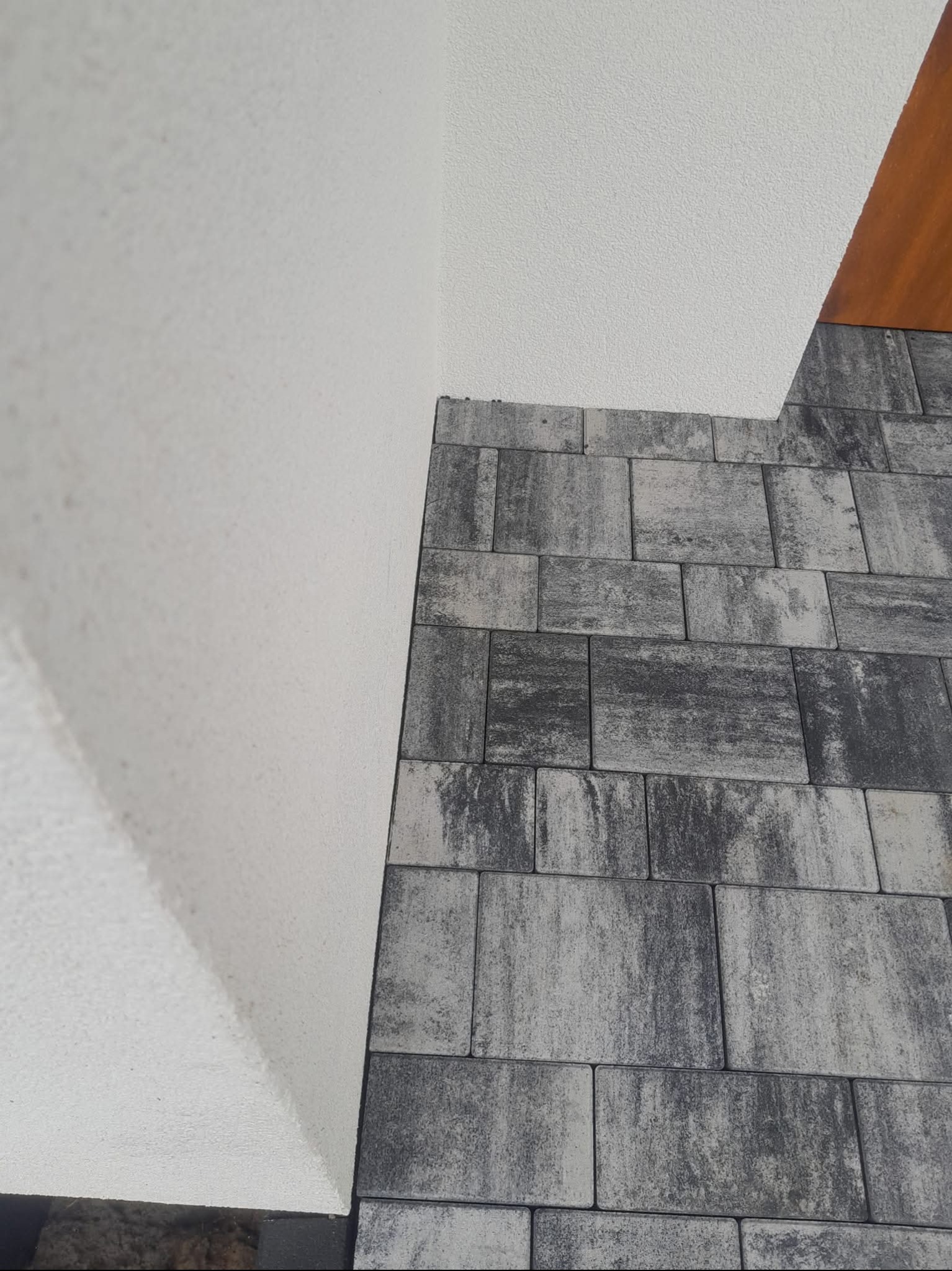 Szara kostka brukowa ułożona przy białej ścianie w Jurgowie. Widoczny fragment drewnianego elementu. Ciekawa faktura kostki i ściany.
