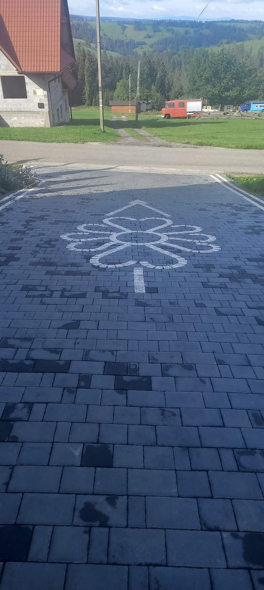 Ułożona, szara kostka brukowa z białym, ozdobnym wzorem przypominającym drzewo. W tle budynek i zalesione wzgórza.
