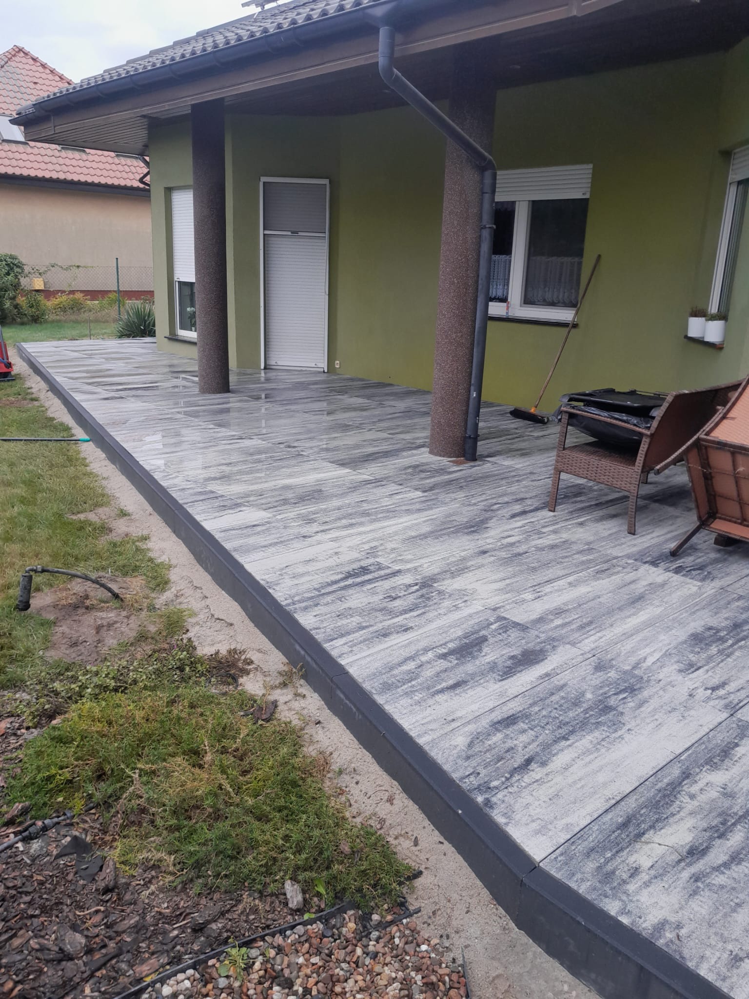 Nowoczesny taras z szarej kostki o nieregularnym wzorze, obrzeża w kolorze antracytowym, widok na zielony dom z kolumnami i częściowo widoczne meble ogrodowe.