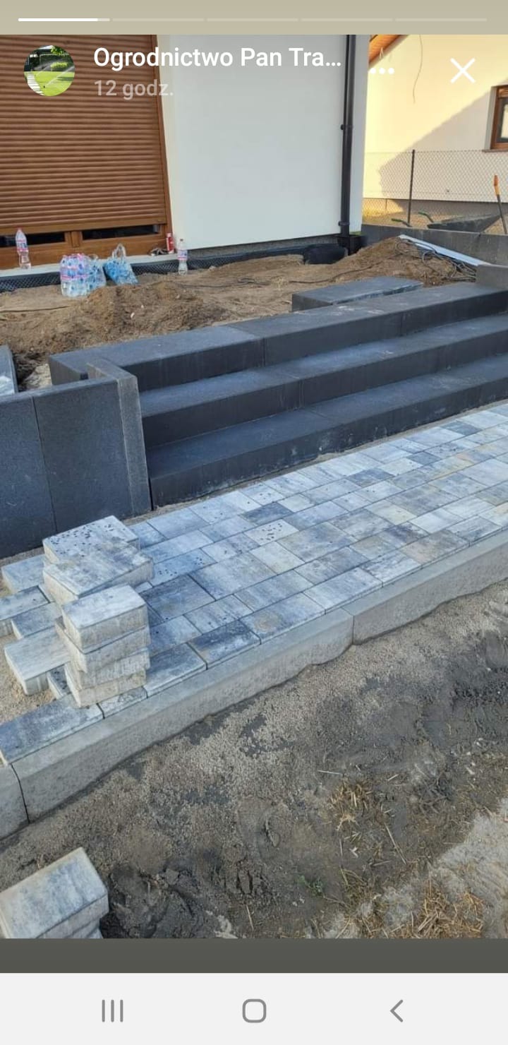 Ułożone schody z ciemnej kostki i fragment chodnika z jasnoszarej kostki. Część kostki ułożona w stos, przygotowana do dalszych prac. Widoczne ślady piasku i ziemi.