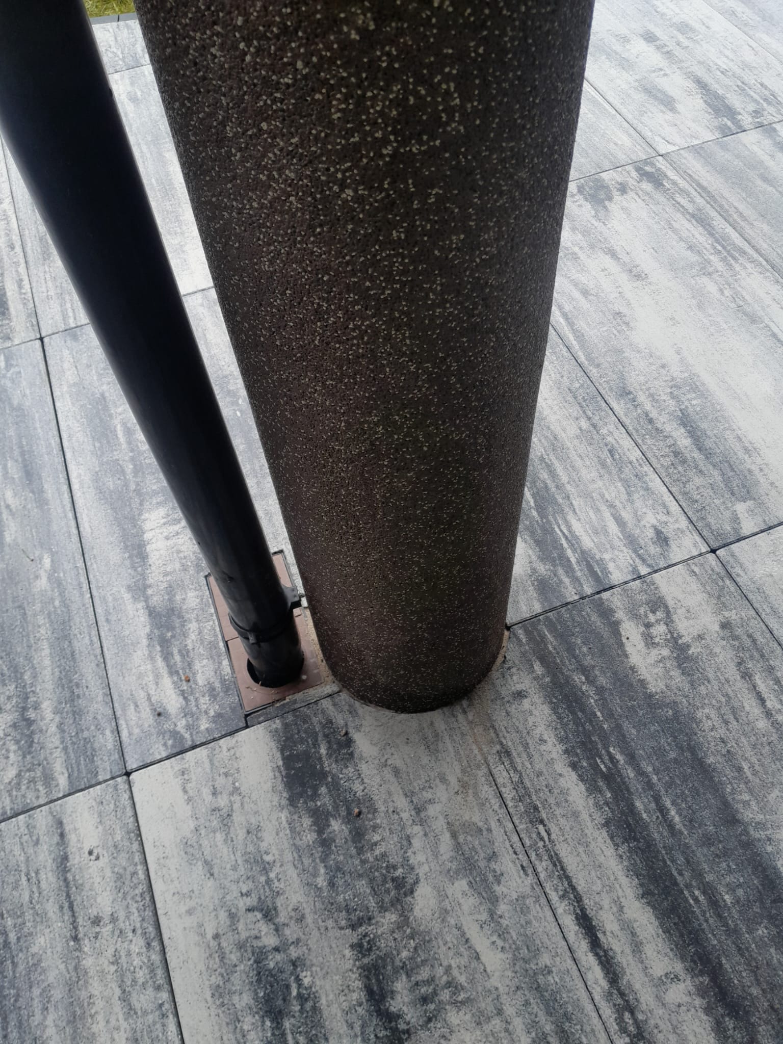 Szara kostka brukowa ułożona wokół czarnej rury i donicy. Widoczny fragment trawnika w tle. Minimalistyczny design nawierzchni.