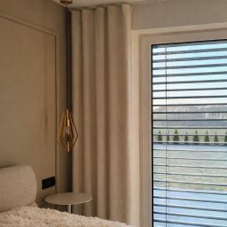 GW Projekt Grzegorz Worobiej - Beżowa zasłona w eleganckim wnętrzu z widokiem na zewnątrz przez żaluzje. Złota lampa wisząca dodaje szyku. Przytulne łóżko z miękką narzutą.
