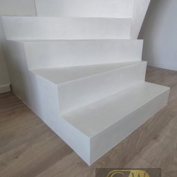 GW Projekt Grzegorz Worobiej - Minimalistyczne, białe schody z mikrocementu, z widoczną strukturą na stopniach, na tle jasnej ściany i drewnopodobnej podłogi. Ujęcie z dołu, podkreślające geometrię.