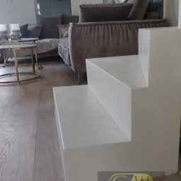 GW Projekt Grzegorz Worobiej - Nowoczesne schody z mikrocementu w jasnym salonie. Minimalistyczny design, drewniana podłoga, szary narożnik i abstrakcyjny obraz w tle. Widoczne logo firmy.